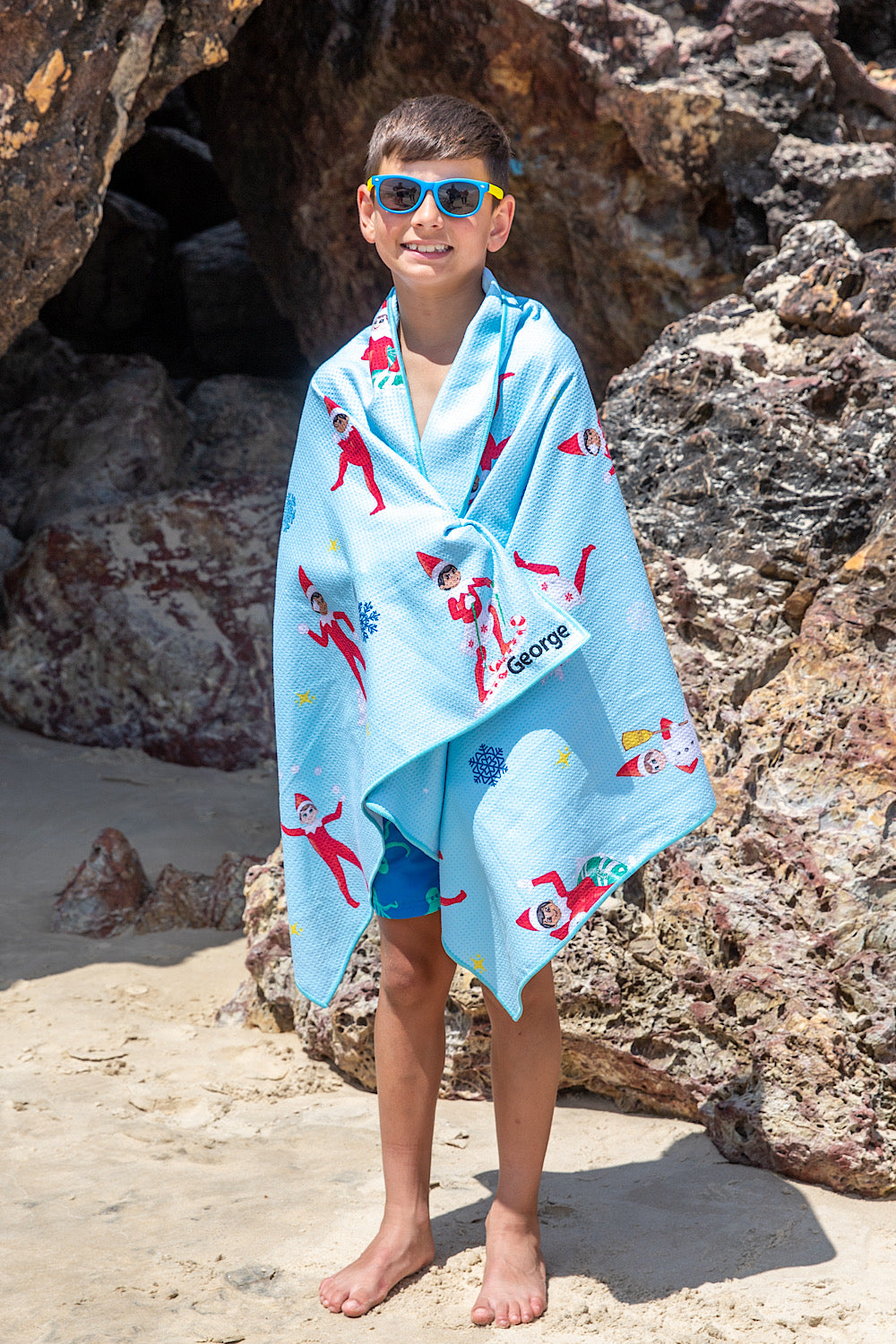 Child wrapped in a blue towel with The Elf On The Shelf characters on a rocky beach