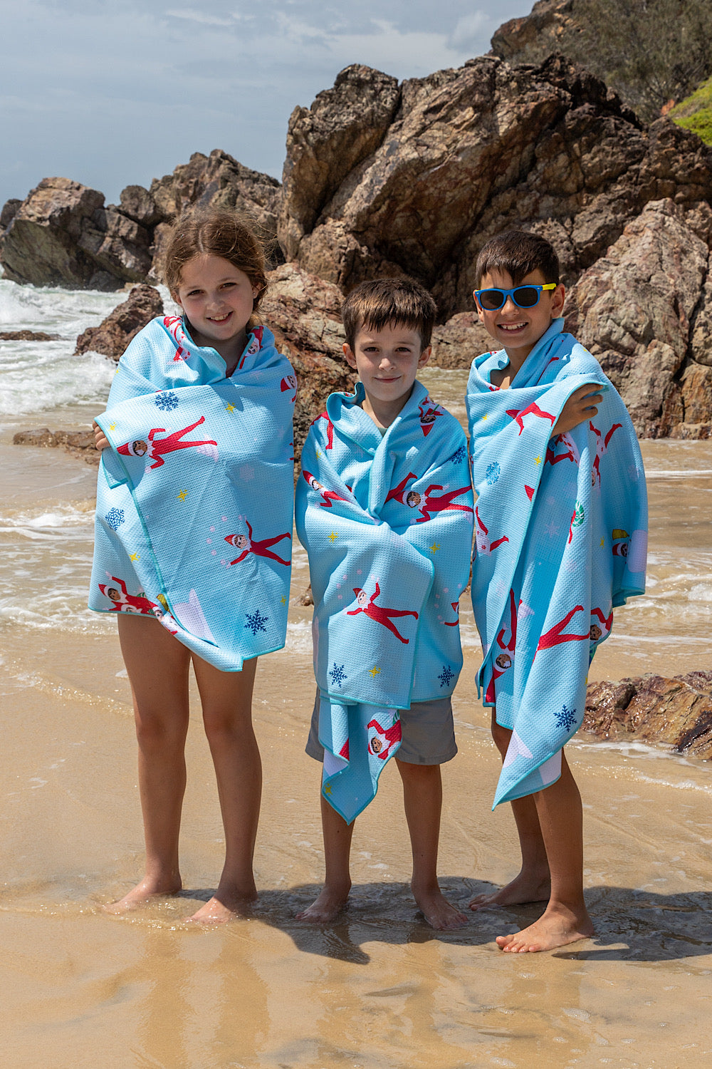 Three children wrapped in blue towels with The Elf On The Shelf patterns on a beach.